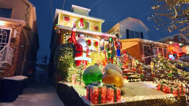 Residential house heavily decorated with Christmas lights and a large Santa. A waving Santa figure, toy soldiers, and large ornaments decorate the snow covered front yard of a home at night.
