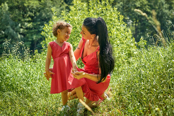 Fototapeta premium A mother and daughter wearing matching red-polka-dot dresses are walking in a summer park. The girl is laughing, her eyes shining with joy and curiosity. Sunlight filters through the tall green grass.