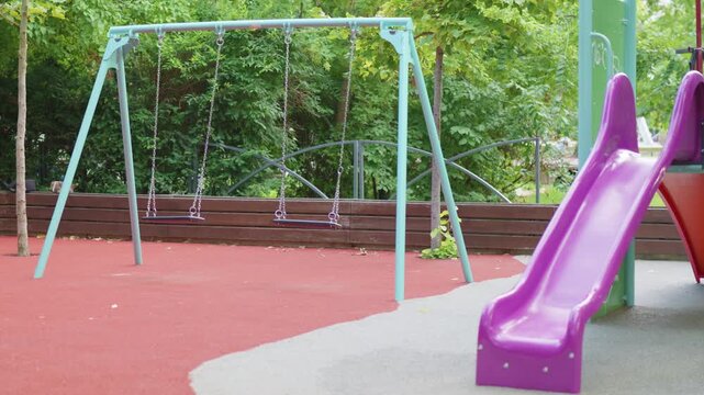 Colorful playground equipment featuring a pink slide and a swing set with empty seats, surrounded by green trees in a vibrant outdoor park setting