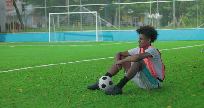 Young Latin Hispanic man of African descent sitting on grass soccer field resting with soccer ball after practice, thoughtful expression near goal net in urban sports park