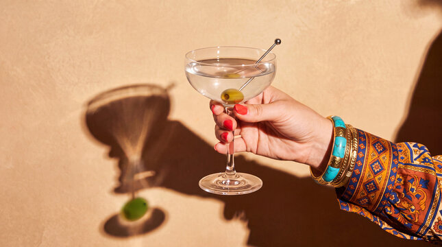 Elegant 1970s Retro Female Hand Holding a Classic Dry Martini Cocktail in Vintage Martini Glass with Olive Garnish, Coral Red Nails, Boho Jewelry and Warm Beige Studio Background