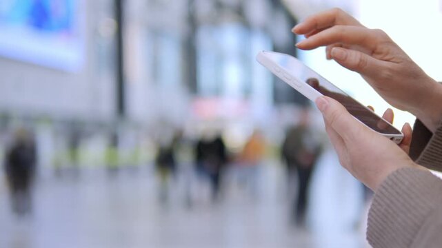 Person holds tablet, fingers tap screen. Blurred background shows busy indoor space. Focus remains on hands and device interaction. Suitable for tech, travel, or digital communication themes