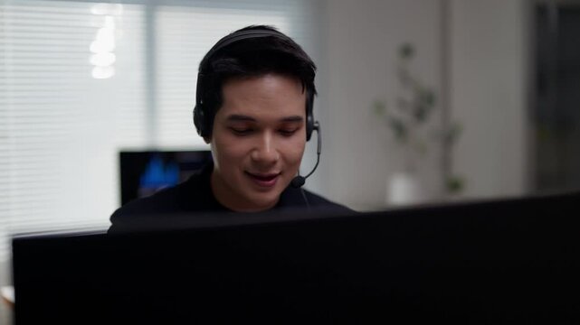 Professional Asian call center agent wearing headset providing friendly customer service and technical support while working in modern office cubicle with computer monitor