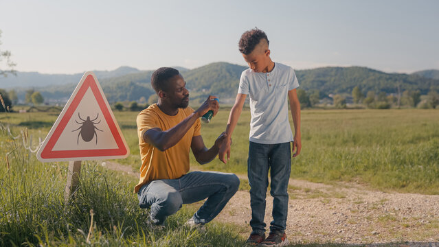 A man sprays tick repellent on his sons arm during an outdoor activity in a grassy field preventing tick bite.