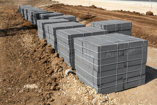 Pallets of grey concrete paving blocks stand in a row on raw dirt ground. The building materials are prepared for sidewalk installation