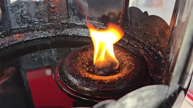 The flame drifts in the wind in an oil lamp, commonly found in temples or shrines. The oil-soaked wick is housed within a donut-shaped holder, with the flame illuminating the space. Fill lamp with oil