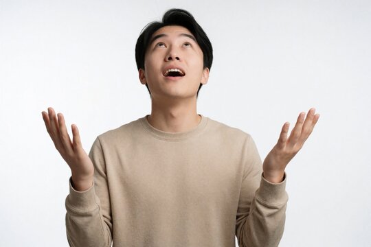 Young man looks up with awe and open palms raised. His mouth is open in wonder, perhaps asking for something or receiving inspiration. He wears simple beige sweatshirt against plain white background.