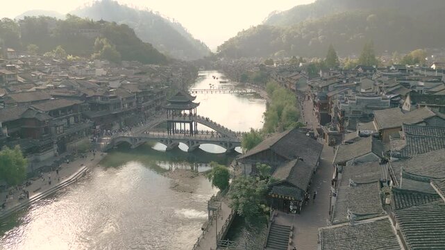 Horizontal 4K UHD aerial drone shot panning left over Tuojiang River and Xueqiao stone bridge surrounded by traditional wooden houses in Fenghuang Ancient Town Hunan China during a hazy day