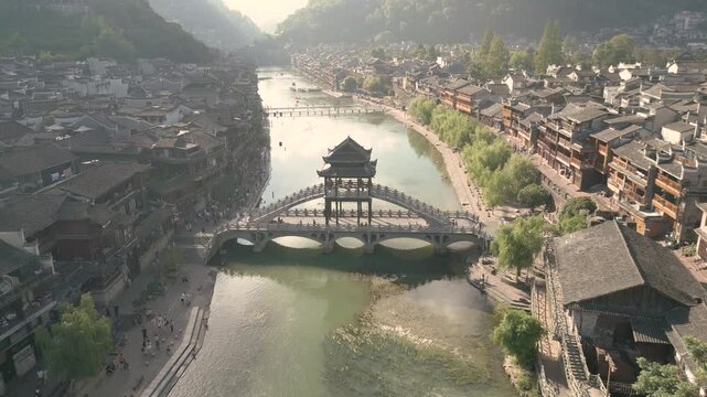 Horizontal 4K UHD aerial drone shot flying forward over Tuojiang River towards Xueqiao Bridge surrounded by traditional wooden houses in historic Fenghuang Ancient Town Hunan China
