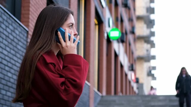 Young woman in red coat holding blue smartphone beside brick city building, side profile beside blurred pedestrian on steps, soft daylight and shallow depth creating urban communication mood
