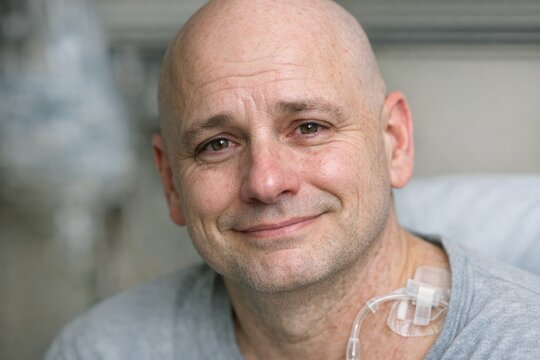 A close-up of a bald man wearing a grey shirt with a plastic tube attached to his neck, used for medical purposes such as a tracheostomy
