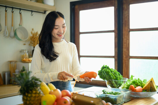 Young Woman Peeling Carrot in Kitchen with Fresh Vegetables and Fruits on Counter