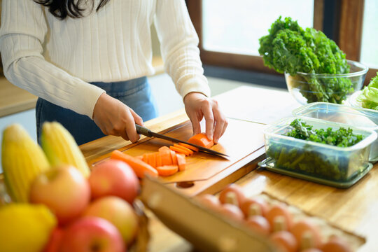Woman Chopping Fresh Carrots on Wooden Cutting Board in Home Kitchen with Vegetables and Fruits