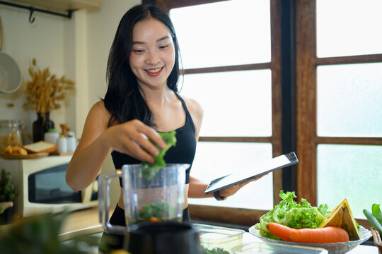 Young Woman Making Kale Smoothie in Kitchen Using Tablet for Recipe, Healthy Lifestyle Wellness Concept