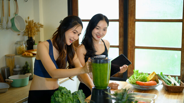 Two Fit Women Making Kale Green Smoothie in Kitchen with Blender and Fresh Vegetables for Healthy Lifestyle