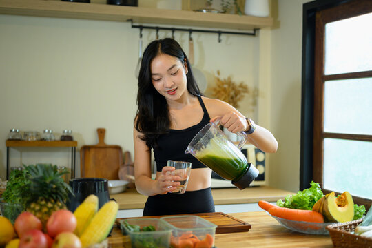 Young Woman Pouring Fresh Kale Smoothie into Glass in Modern Kitchen Surrounded by Fruits and Vegetables
