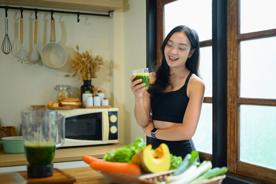 Young Woman Holding Green Kale Smoothie in Kitchen Surrounded by Fresh Vegetables for Healthy Lifestyle