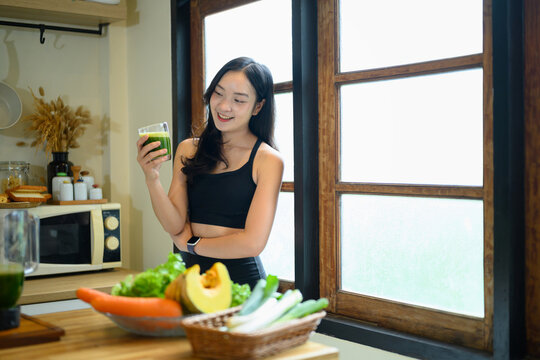 Young Woman in Sportswear Holding Green Kale Smoothie in Kitchen with Fresh Vegetables for Healthy Lifestyle