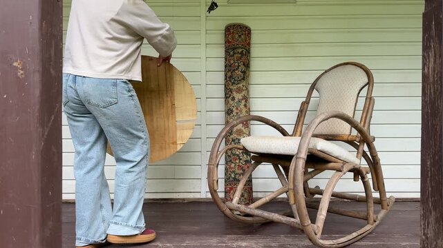 A woman carries a rolled vintage rug and rearranges furniture on a country house veranda during an outdoor makeover. Rustic rocking chair, round wooden table, and cozy porch decor create a warm DIY 