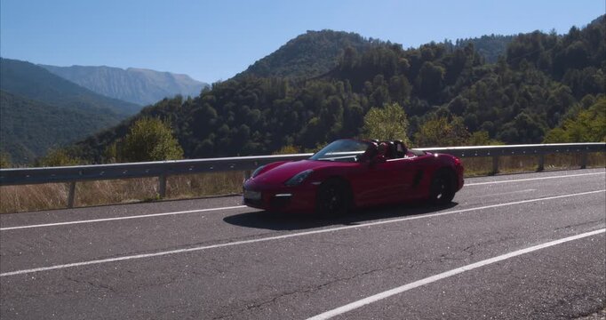 Red convertible passes in profile above a broad forested mountain