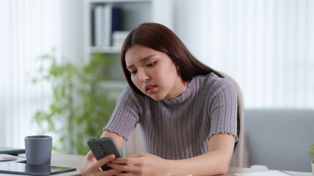 Young Asian woman looking concerned at smartphone with frustrated expression in modern home office