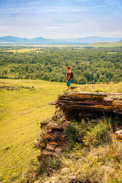 Hiker with backpack stands on edge of mossy cliff, admiring endless panorama of forest-steppe with mountains on horizon. A solo traveler without face, looking into distance, slow travel