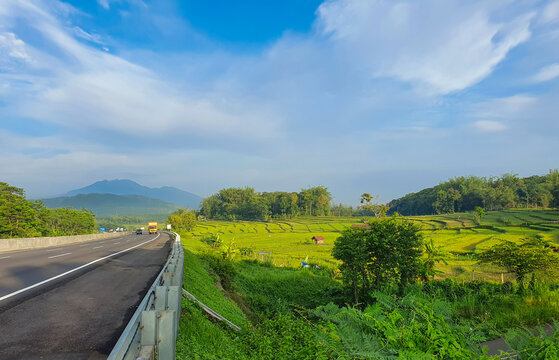Scenic toll road beside lush green rice fields in Central Java, Indonesia, under a dramatic sky. Ideal for travel, infrastructure, and distribution.