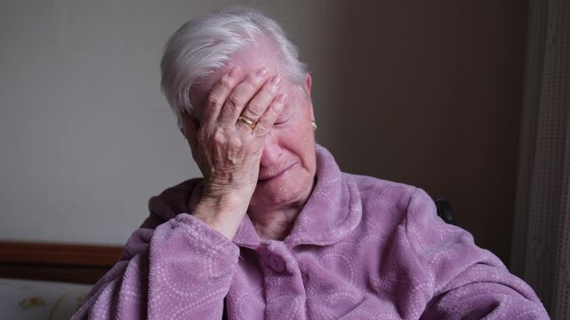 Desperate senior woman with white hair sitting indoors, covering her face with her hand in a gesture of grief, sadness, and despair, expressing loneliness and mental health problems