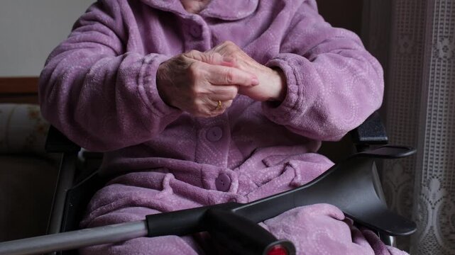 Worried senior woman with mobility issues sitting in a wheelchair, anxiously rubbing her wrinkled hands together with a crutch resting on her lap, showing concern and loneliness