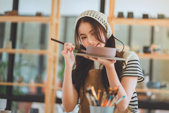 Asian woman artist painting handmade ceramic product in pottery studio &ndash; creative process of decorating sculpture with brush for small business, art workshop, and modern craft design startup