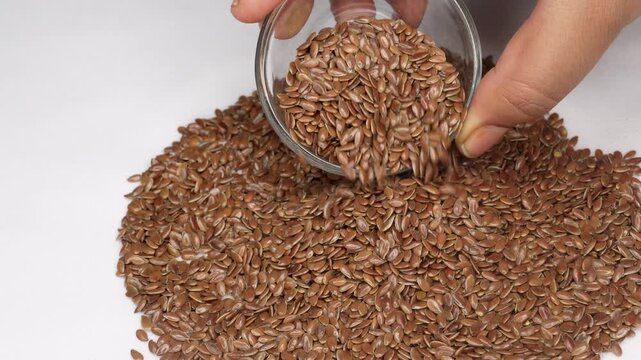 Woman hand glass bowl to pouring of flaxseeds with heap of dry brown flax seeds, isolated on white background. healthy food ingredient for omega-3s concept.