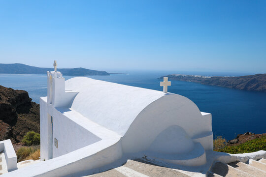 A serene white chapel with crosses stands above the deep blue Aegean Sea, capturing the timeless beauty and spiritual calm of Santorini&rsquo;s cliffside architecture under a clear summer sky.