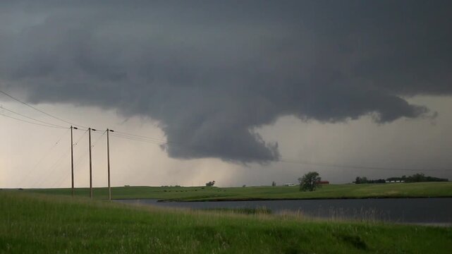 A mostly invisible tornado churns dirt underneath a wall cloud as a dangerous storm passes through a rural area.