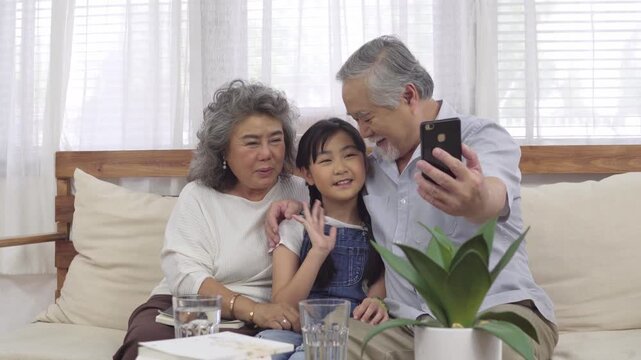 Asian multi generation family sitting together on couch taking selfie photo with smartphone at home happy grandmother grandfather and granddaughter waving hand smiling making video call