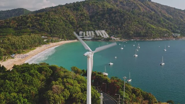 Phuket, Nai Harn Beach, aerial wind turbine generating renewable electricity on a lush hilly tropical coastline overlooking boats moored in the turquoise bay and tourist umbrellas on the sandy shore