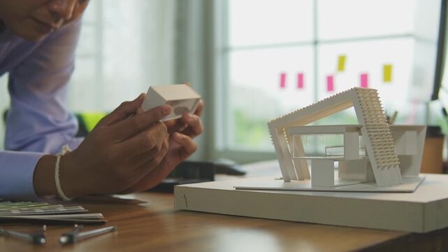 Asian male architect holding examining small architectural scale model in creative office. concept of architectural design, construction planning building structure engineering and real estate.
