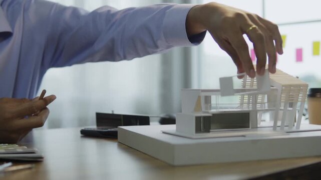 Asian male architect holding pencil and placing building section on architectural scale model at wooden desk in office. concept of architectural design and construction planning building structure.