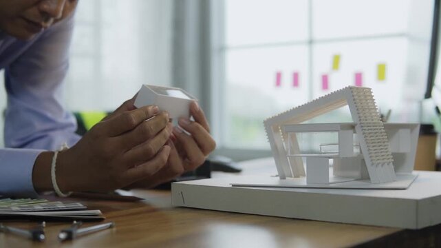 Asian male architect holding examining small architectural scale model in creative office. concept of architectural design, construction planning building structure engineering and real estate.