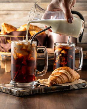 Pouring milk into a glass of iced coffee with another in behind and a croissant.