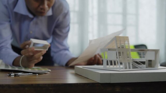Asian male architect holding blueprint paper and examining small building section on architectural scale model in office. concept of architectural design and construction building structure design.
