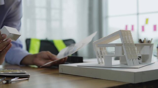 Asian male architect holding blueprint paper and examining small building section on architectural scale model in office. concept of architectural design and construction building structure design.