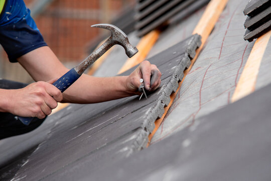 Worker installs roofing tiles on a building during daylight hours using a hammer and nails for construction work