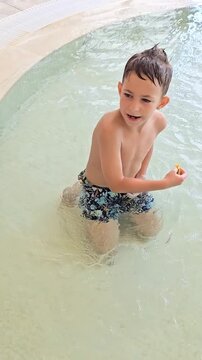Six year old boy playing in a shallow children's splash pool with colorful beach toys, bucket and sand molds. Happy summer water play, family vacation, outdoor fun and carefree childhood