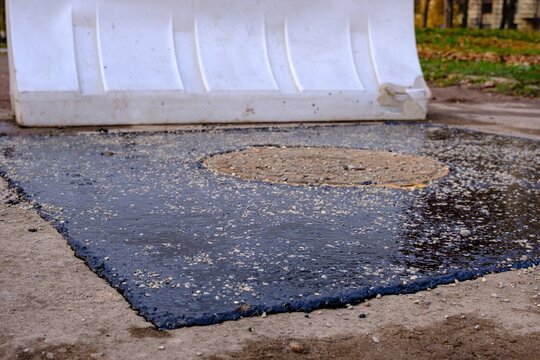 A freshly patched section of wet black asphalt surrounds a circular rusty manhole cover, with light gravel scattered across the glossy surface and a white plastic barrier in the blurred background