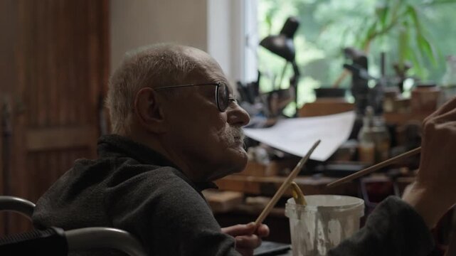 Senior artist on a wheelchair paints an oil painting on canvas in a studio