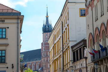 Kraków – Stare Miasto w oddali, Kościół Mariacki. Widok z miejskiej ulicy pełnej zabytkowych budynków. Gotycka wieża kościoła z misterną iglicą wyraźnie wznosi się na tle błękitnego nieba.  © Grzegorz