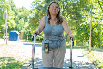 Obraz premium Middle-aged woman walking rollator paved path, mobility aid assistance and independence, Rehabilitation therapy exercises outdoors, activity disabled person moving independently, physical limitations