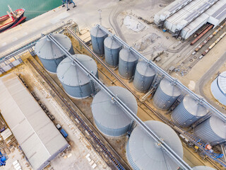 Grain silos stand at a shipping port where cargo ships load and unload materials during daylight hours © DSMT