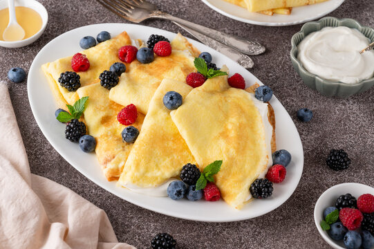 Crepes or thin pancakes with cream cheese, fresh berries and honey on a white plate on a concrete background. Copy space