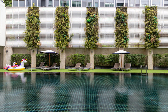 A scenic swimming pool area featuring dark tiled water, multiple sun loungers, umbrellas, a unicorn inflatable float, artificial turf, and a striking vertical garden wall with white slatted panels.